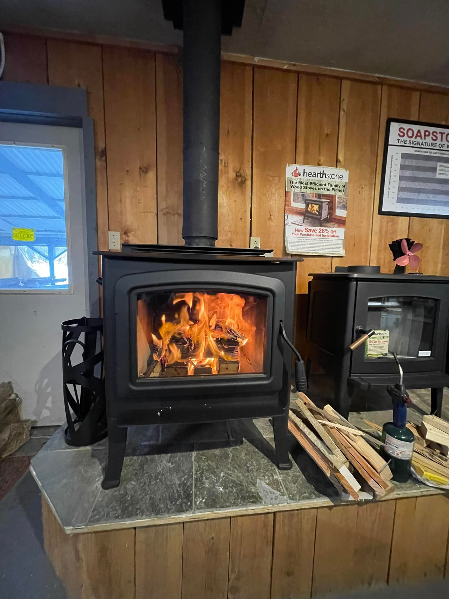 Wood-burning stove with flames, chimney, and wood logs inside a room with wooden walls.