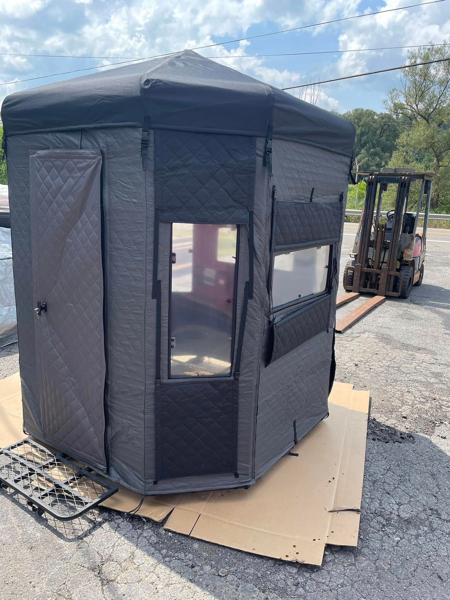 Hexagonal, gray hunting blind with windows, door, and black roof, on cardboard. A forklift is visible in the background.
