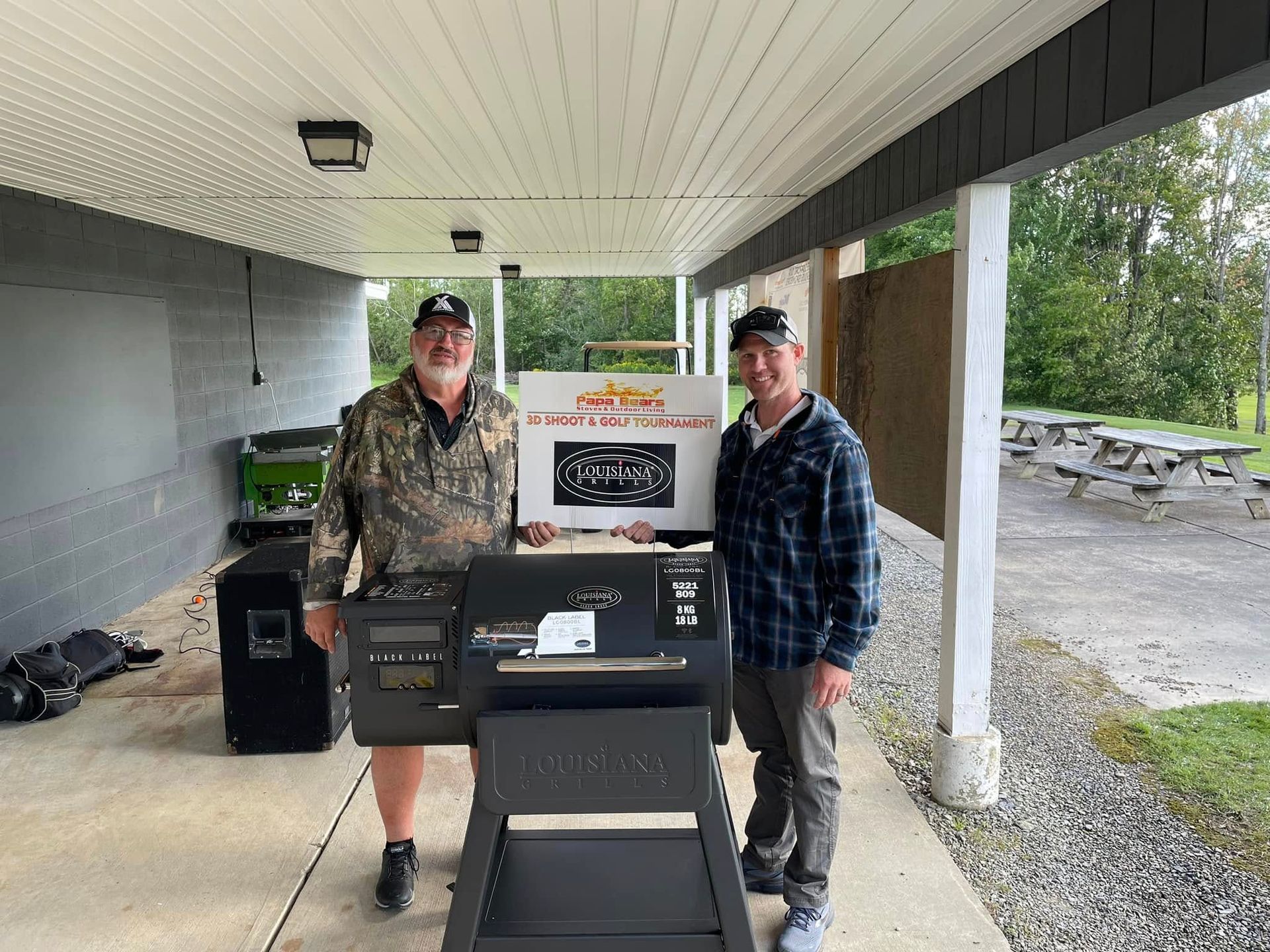 Two men stand with a smoker grill and a sign, under a covered area, outdoors.