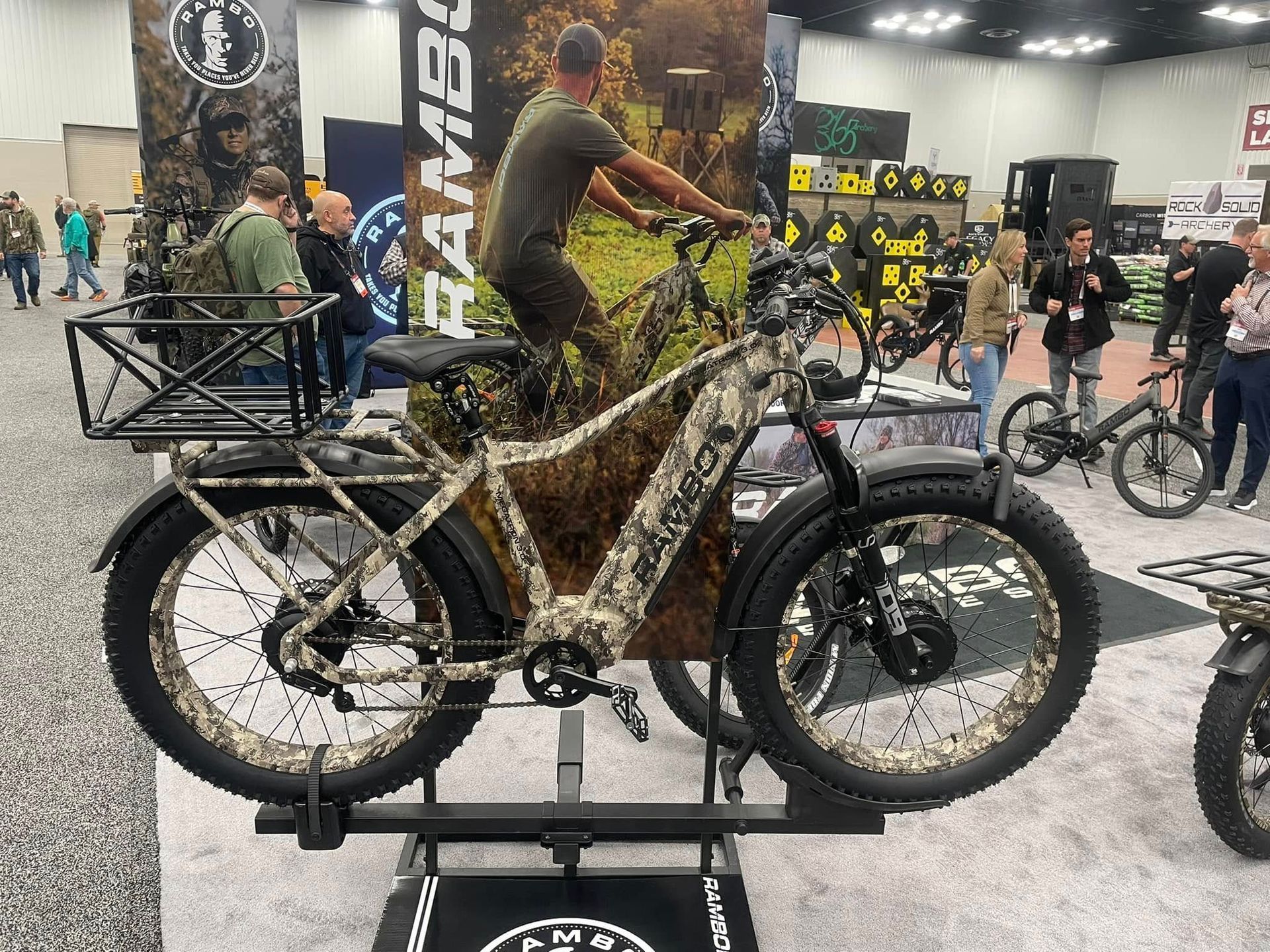 Camouflage electric bike on display at an event, with rider graphic in background.