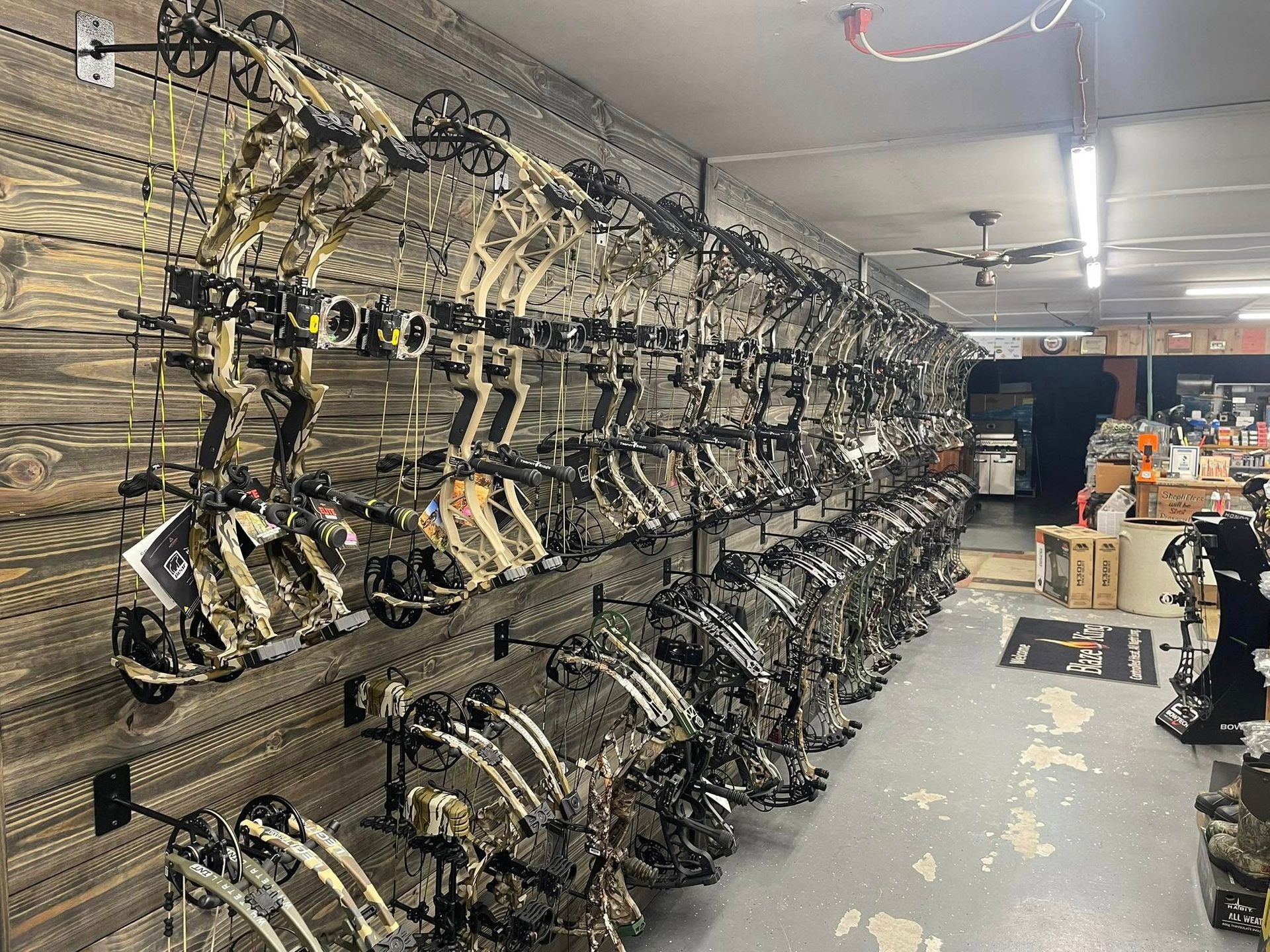 A wall displaying many camouflage compound bows inside a store, with other hunting supplies visible.