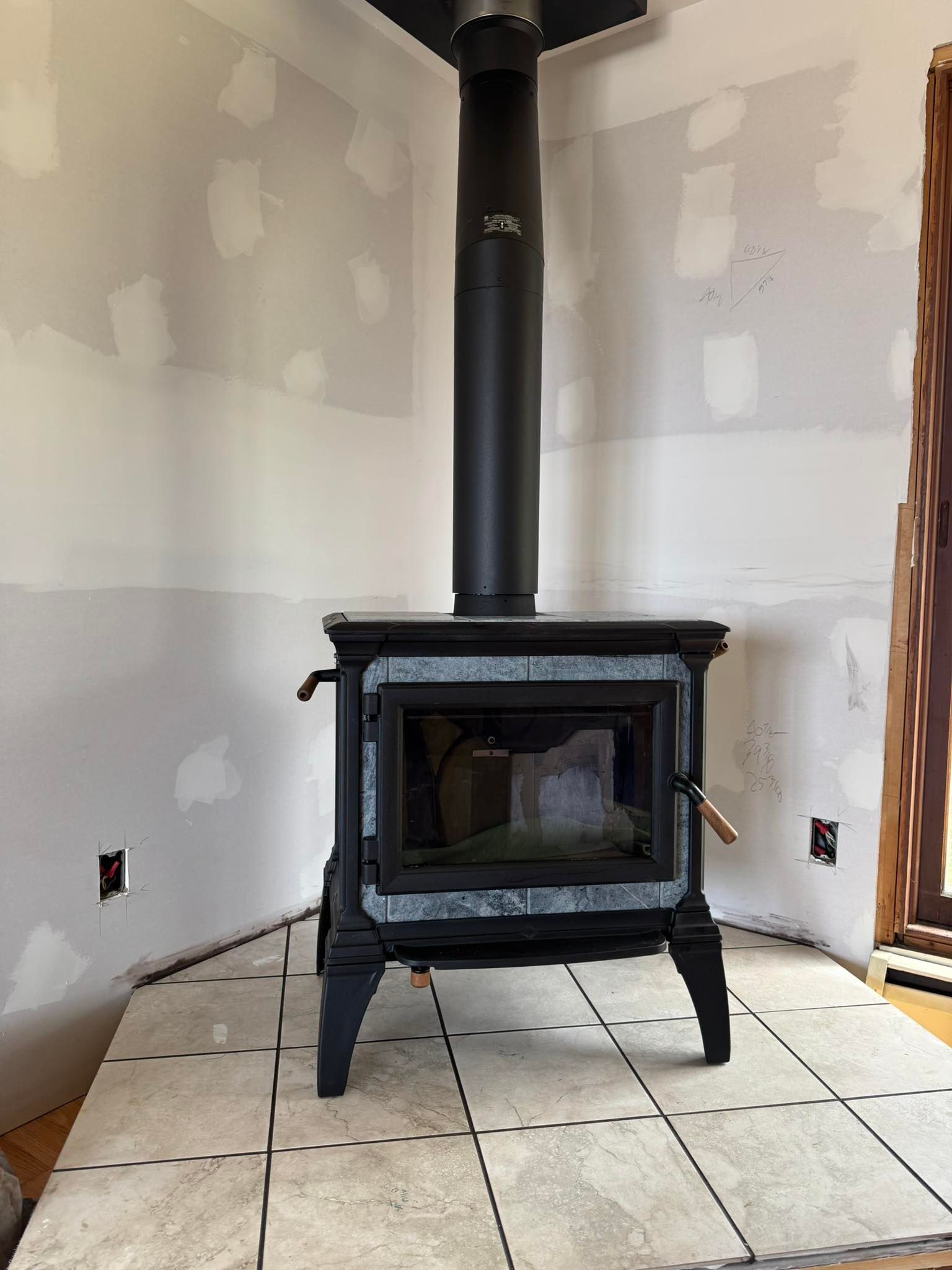 Black wood-burning stove with black chimney pipe, on tiled floor in corner of room, drywall walls.