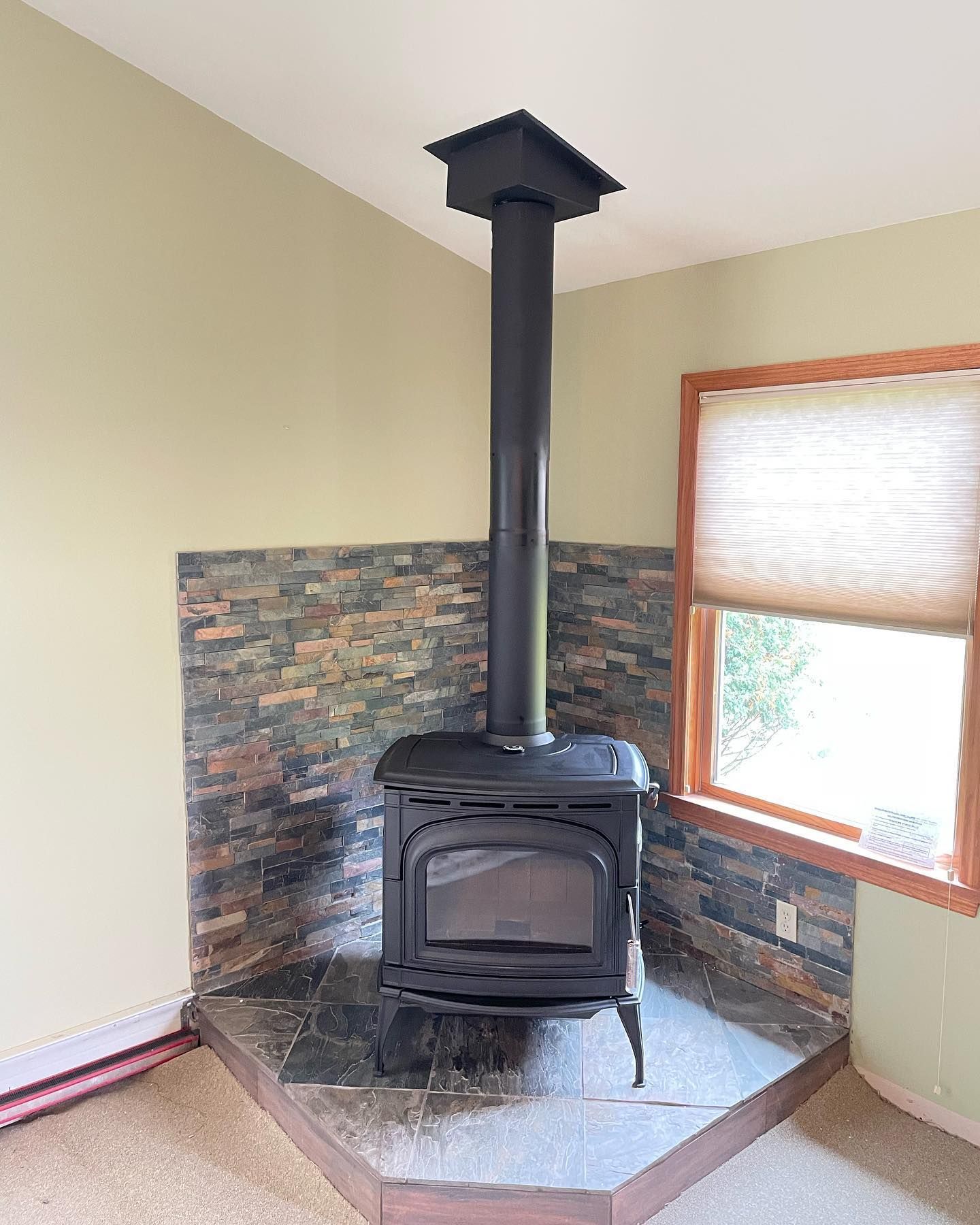 Black wood-burning stove with chimney in corner of a room, against a stone tiled wall.