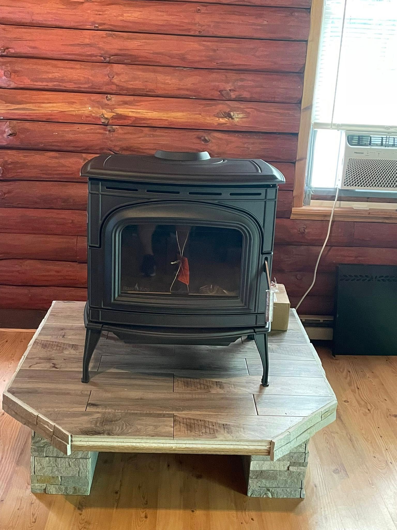 Black wood-burning stove on a wood and brick hearth against a red log wall and window.
