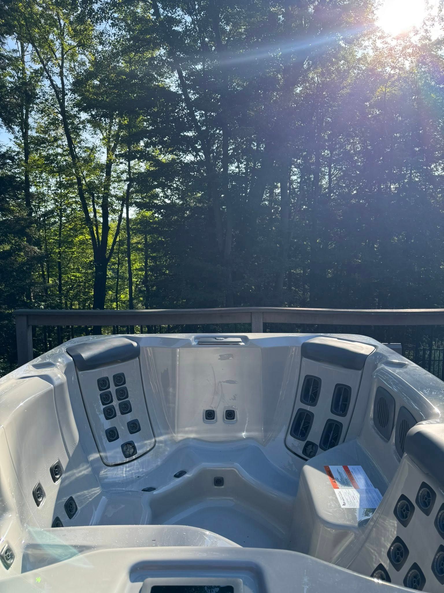 Hot tub on a deck, backlit by sun. Gray tub with multiple jets, controls, and forest view.