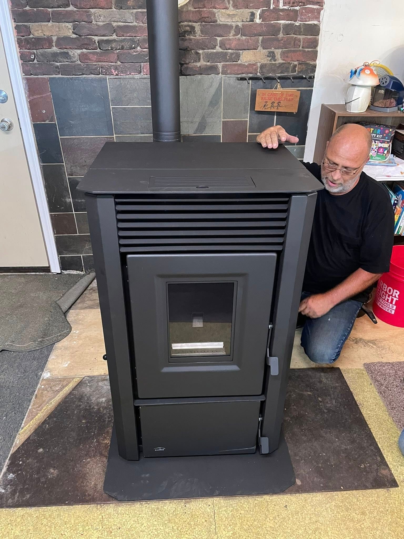 Man kneeling beside a black pellet stove, touching the top, indoors with brick wall.
