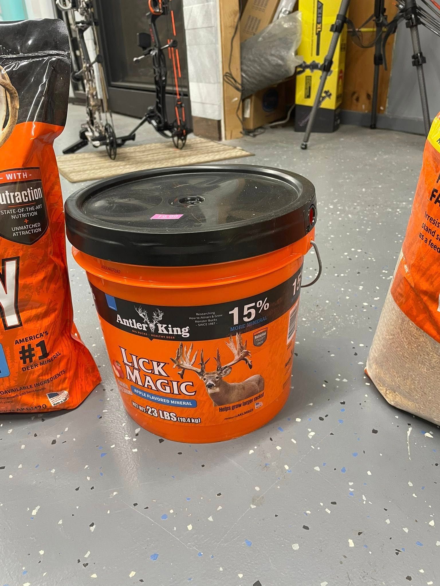 Orange bucket with black lid, deer feed, and bags of feed on a garage floor.