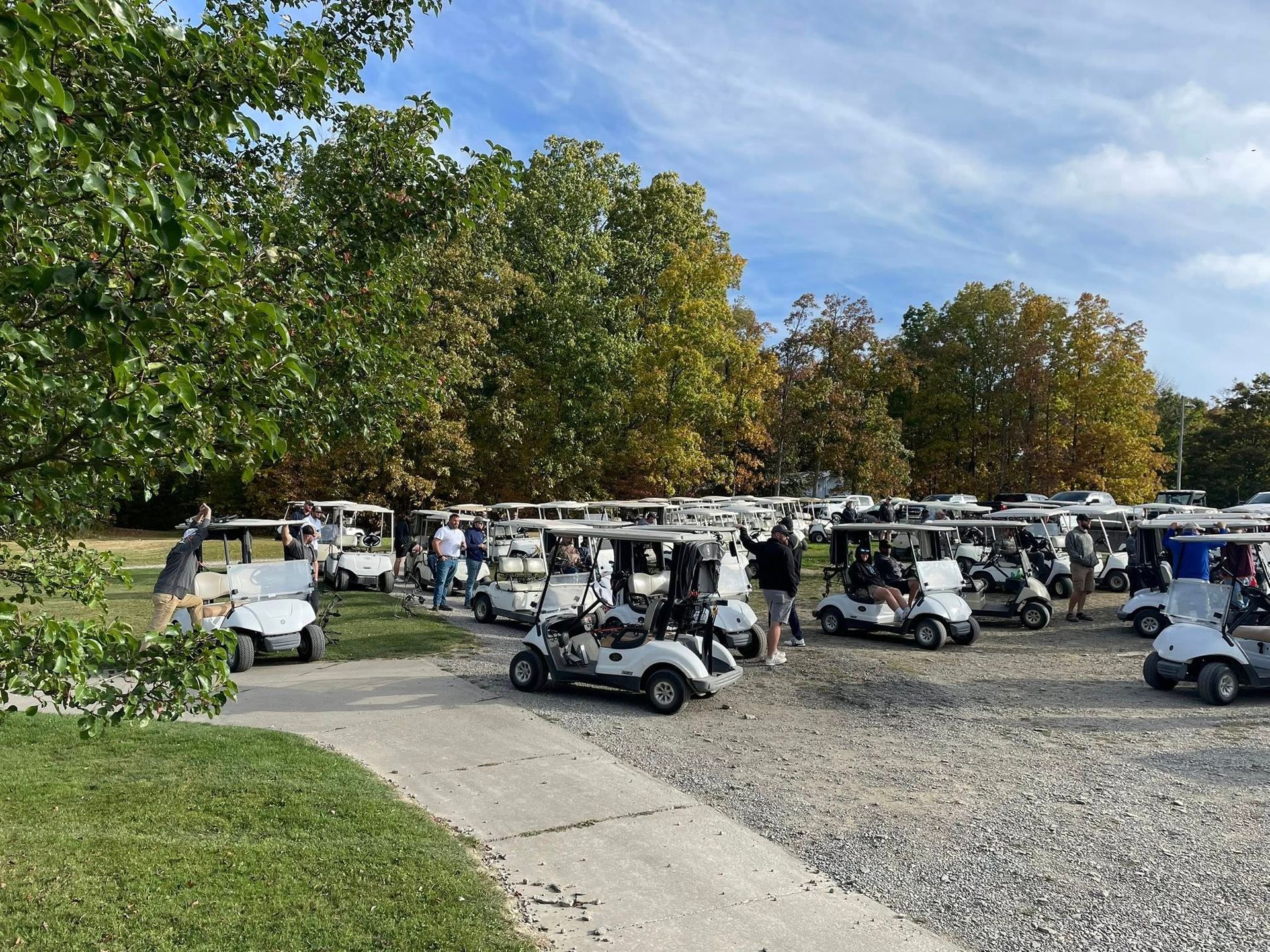 Golf carts parked on gravel near a green grassy area and trees under a cloudy sky.