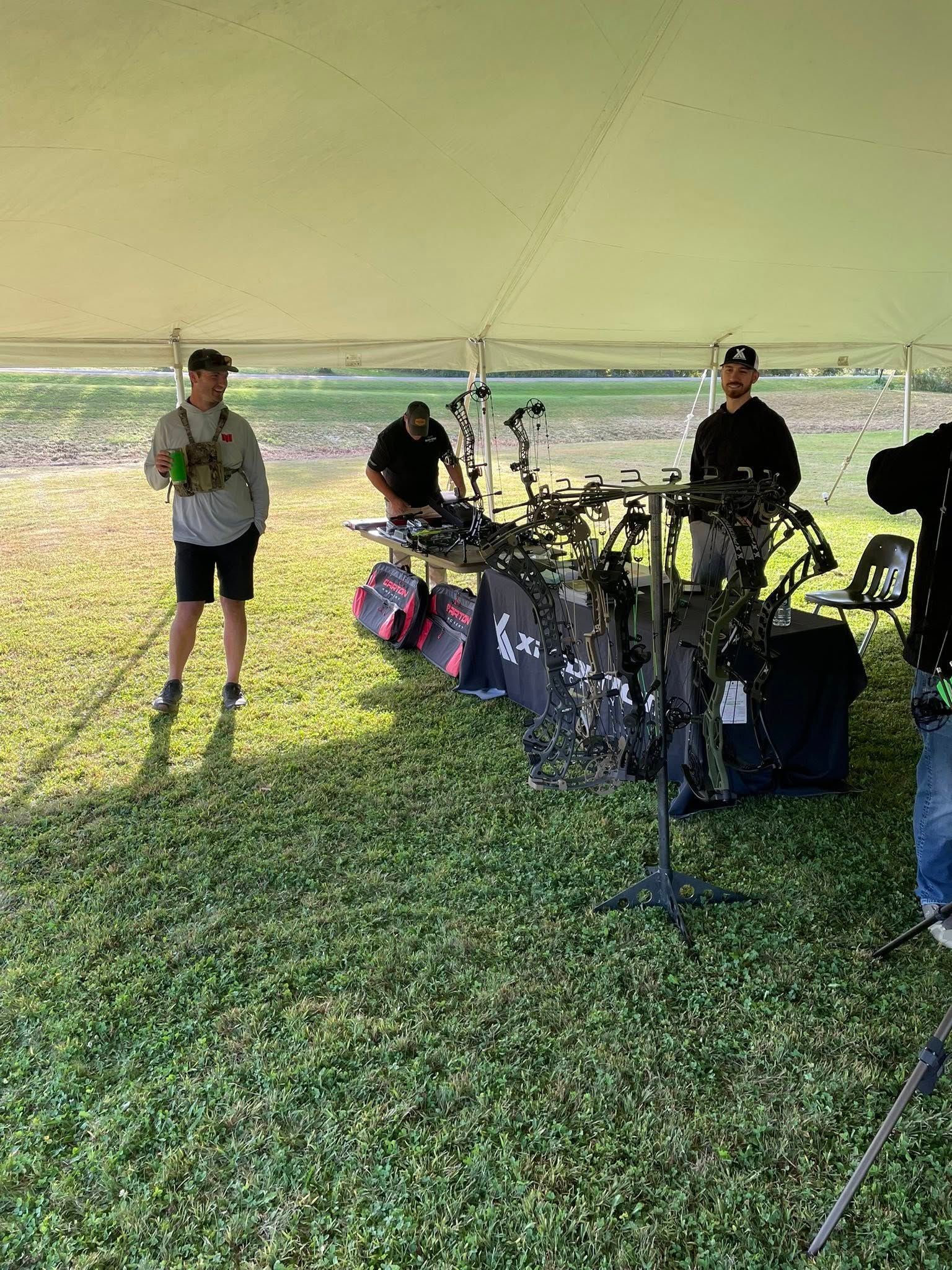 People under a tent, looking at compound bows set up on a table. Archery competition or display.