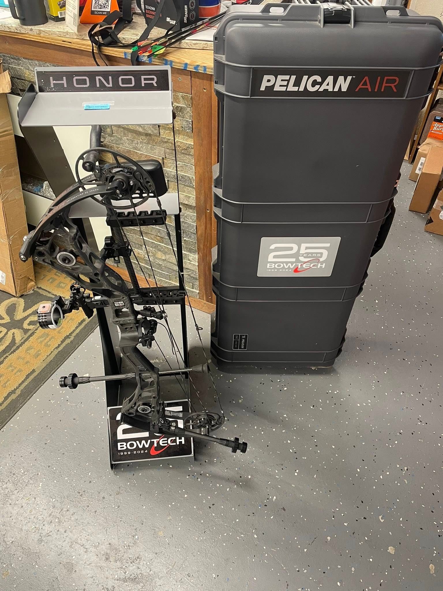 Compound bow on a stand next to a black Pelican Air case. Indoors, gray flooring.