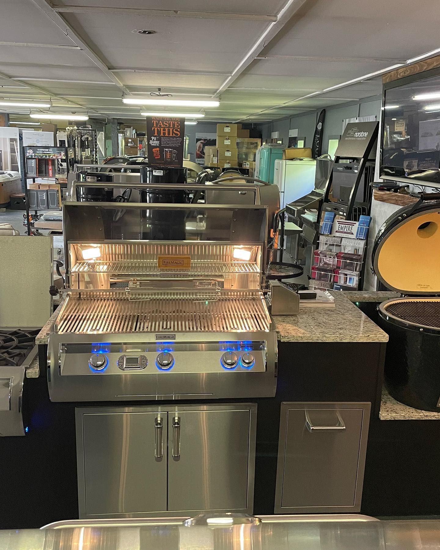 Stainless steel grill with blue lit knobs in a showroom, with other appliances in the background.