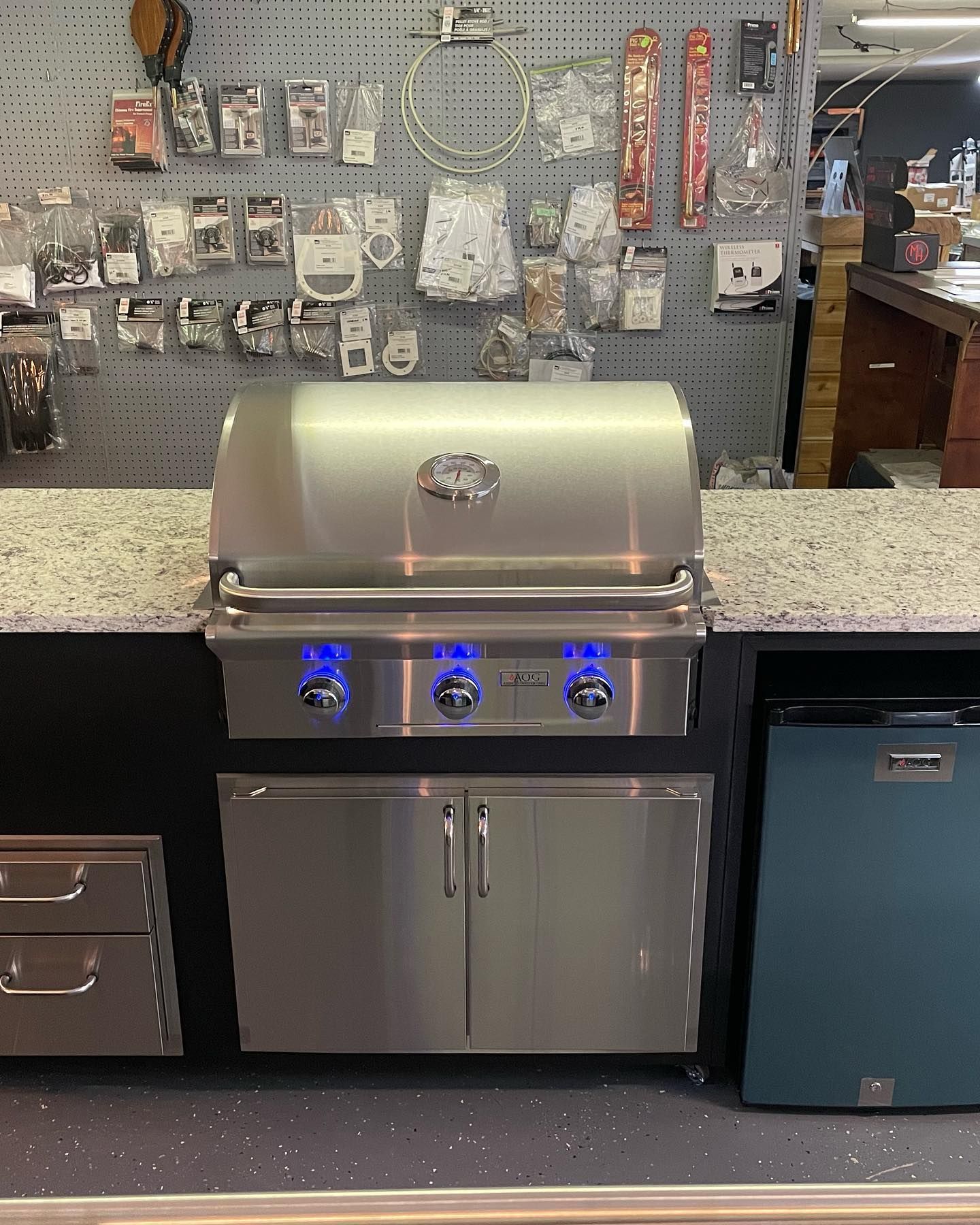 Stainless steel outdoor grill with blue-lit knobs, built into a counter with a cabinet and small refrigerator.