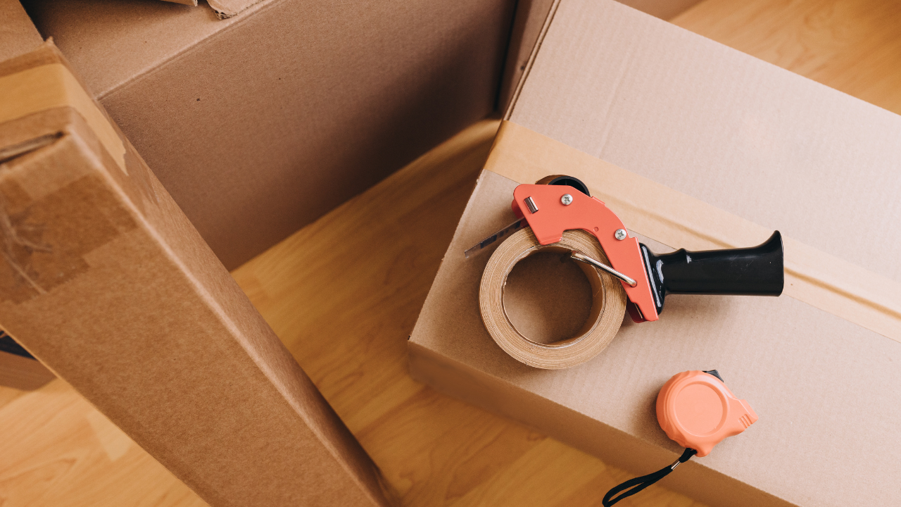 Cardboard boxes, tape dispenser, and measuring tape on a wooden floor, suggesting packing or moving.