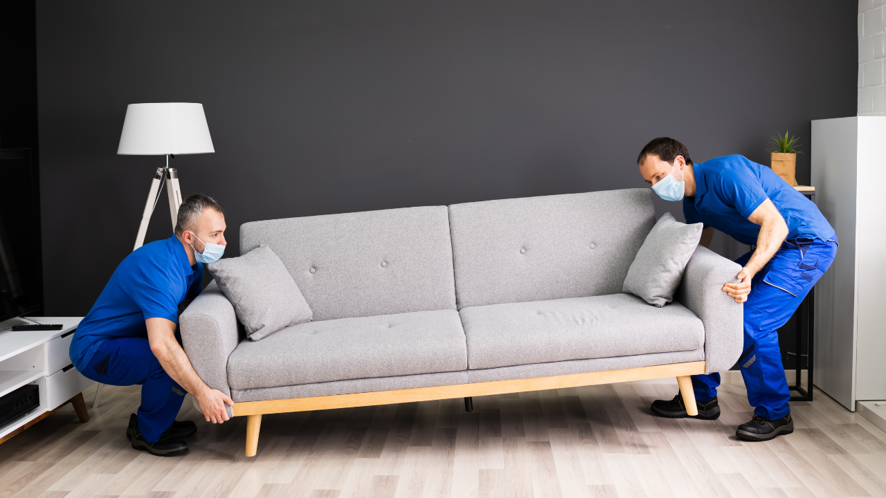 Two movers in blue coveralls and masks lift a grey couch in a room with a lamp and dark gray wall.