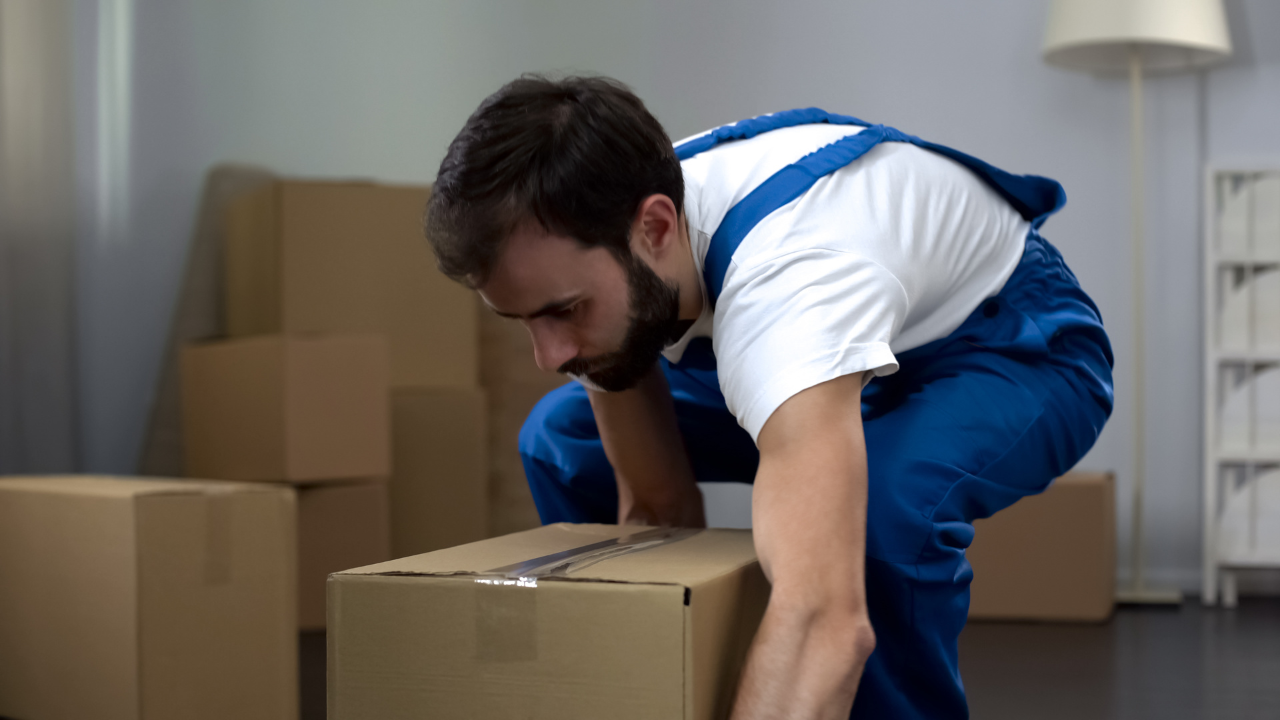 Man in blue overalls lifting a cardboard box indoors, other boxes in background.