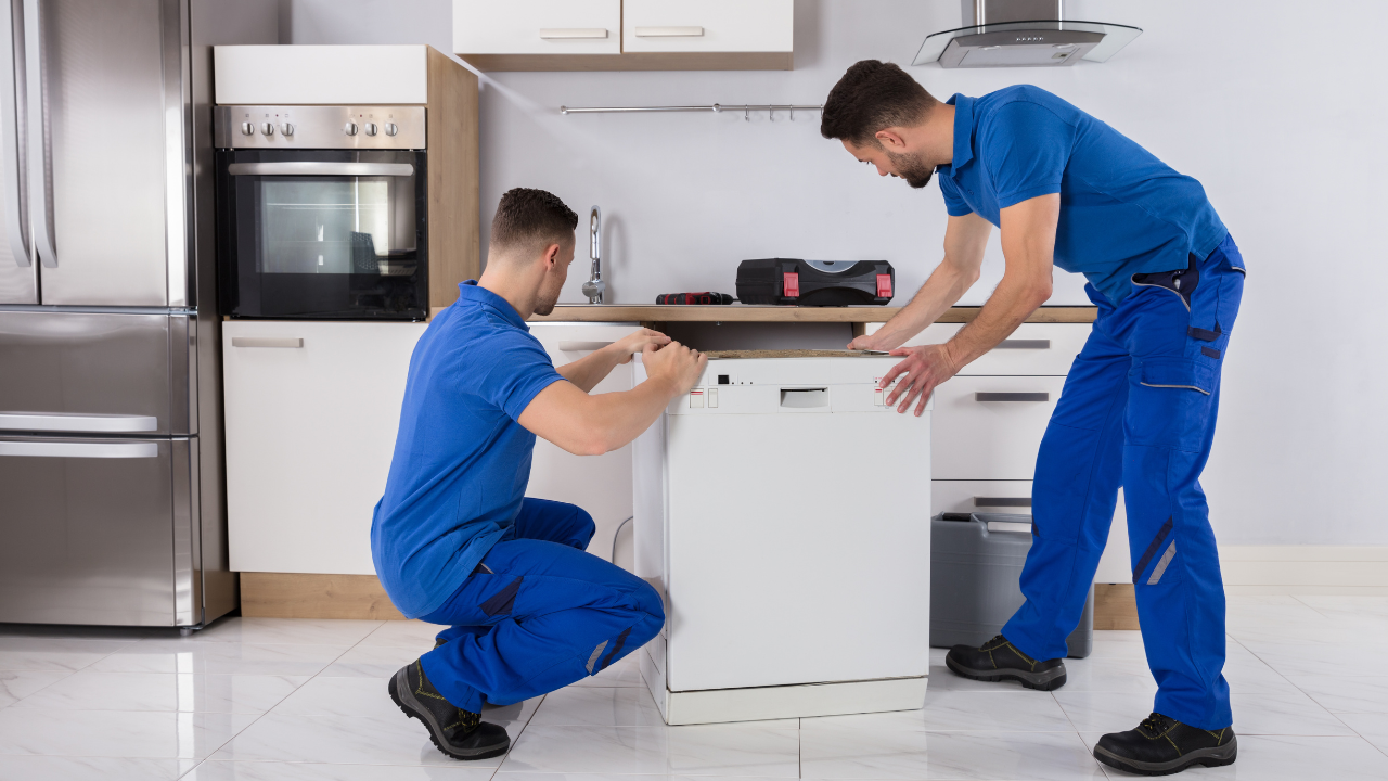 Two repairmen in blue uniforms installing a dishwasher in a kitchen.