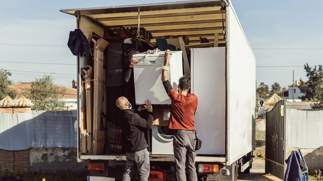 Two people loading a white appliance into a moving truck. Sunny outdoor setting.