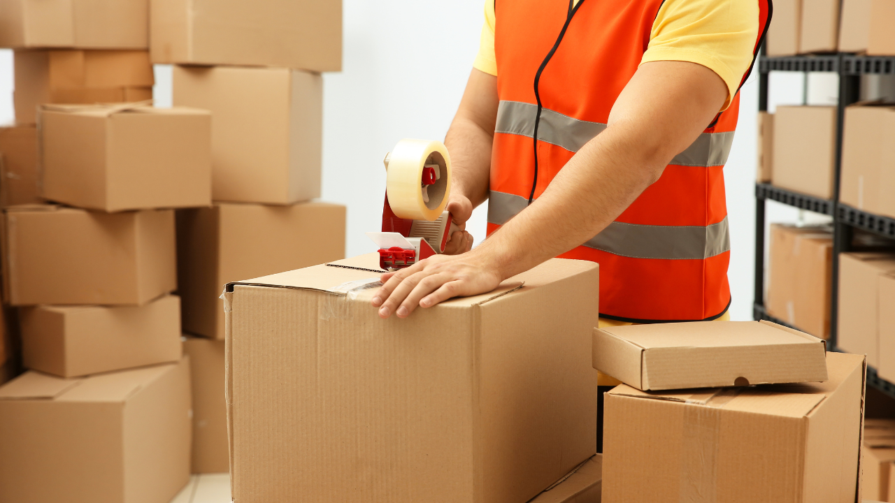 Worker in orange vest taping a cardboard box in a warehouse, surrounded by stacks of packages.