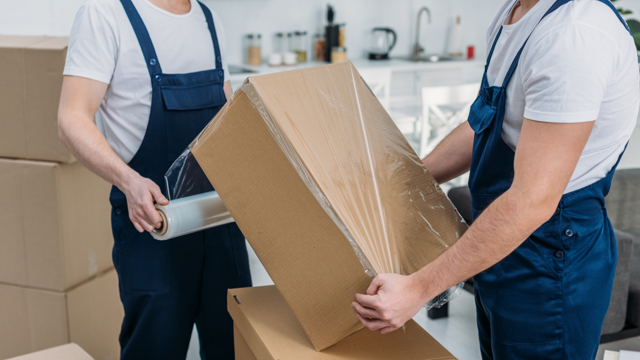 Two movers wrapping a cardboard box in plastic wrap, inside a home.