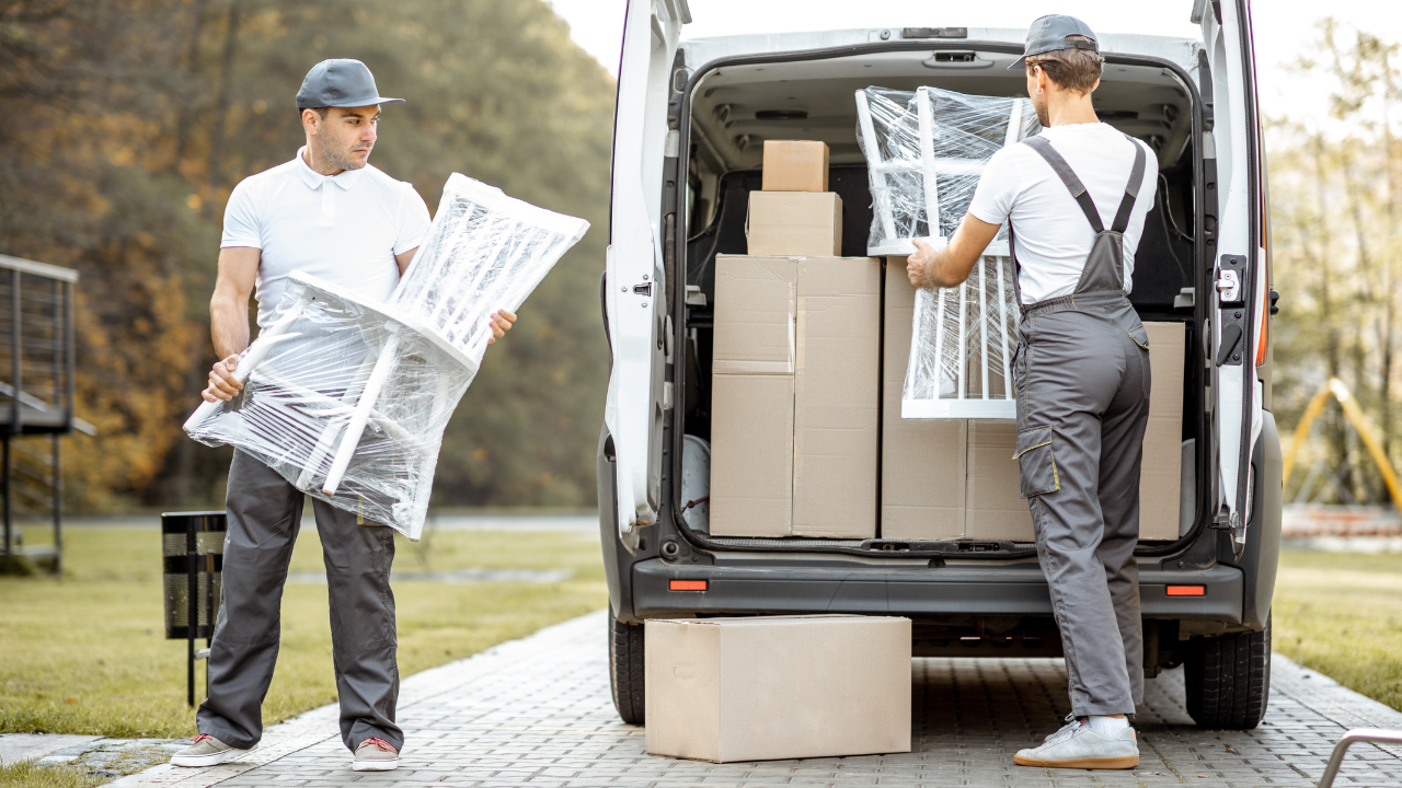 Two movers loading a van with wrapped furniture and boxes outdoors.