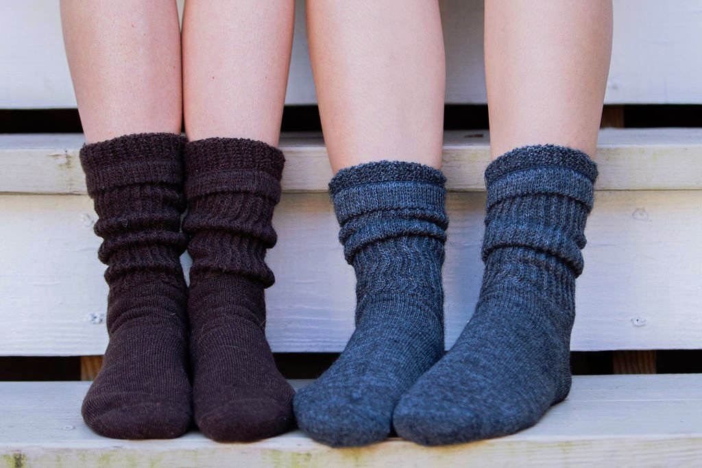 Two pairs of legs, each wearing a different color knitted sock. Brown and gray socks on a wooden bench.