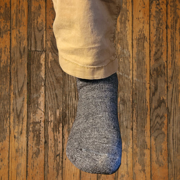 Person's legs wearing tan pants and a gray sock. Standing on a brown, possibly wood, surface.