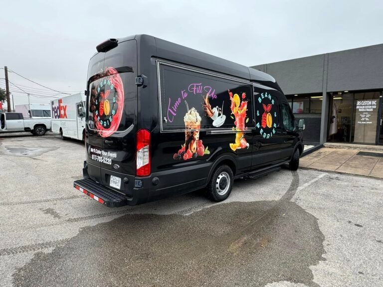 Black food truck with colorful food graphics parked in a lot outside a commercial building.