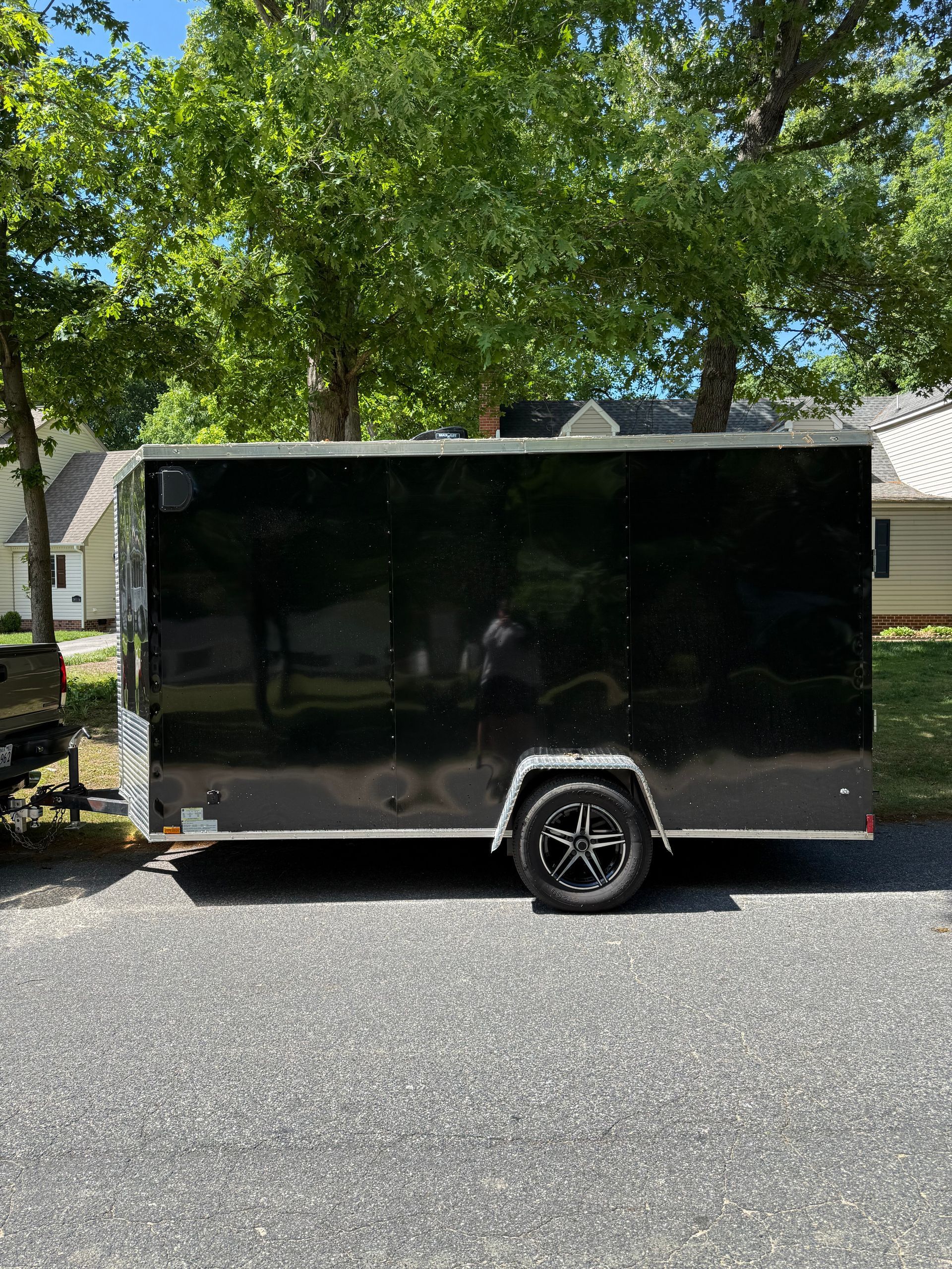 Black enclosed utility trailer parked on a residential street under leafy trees.