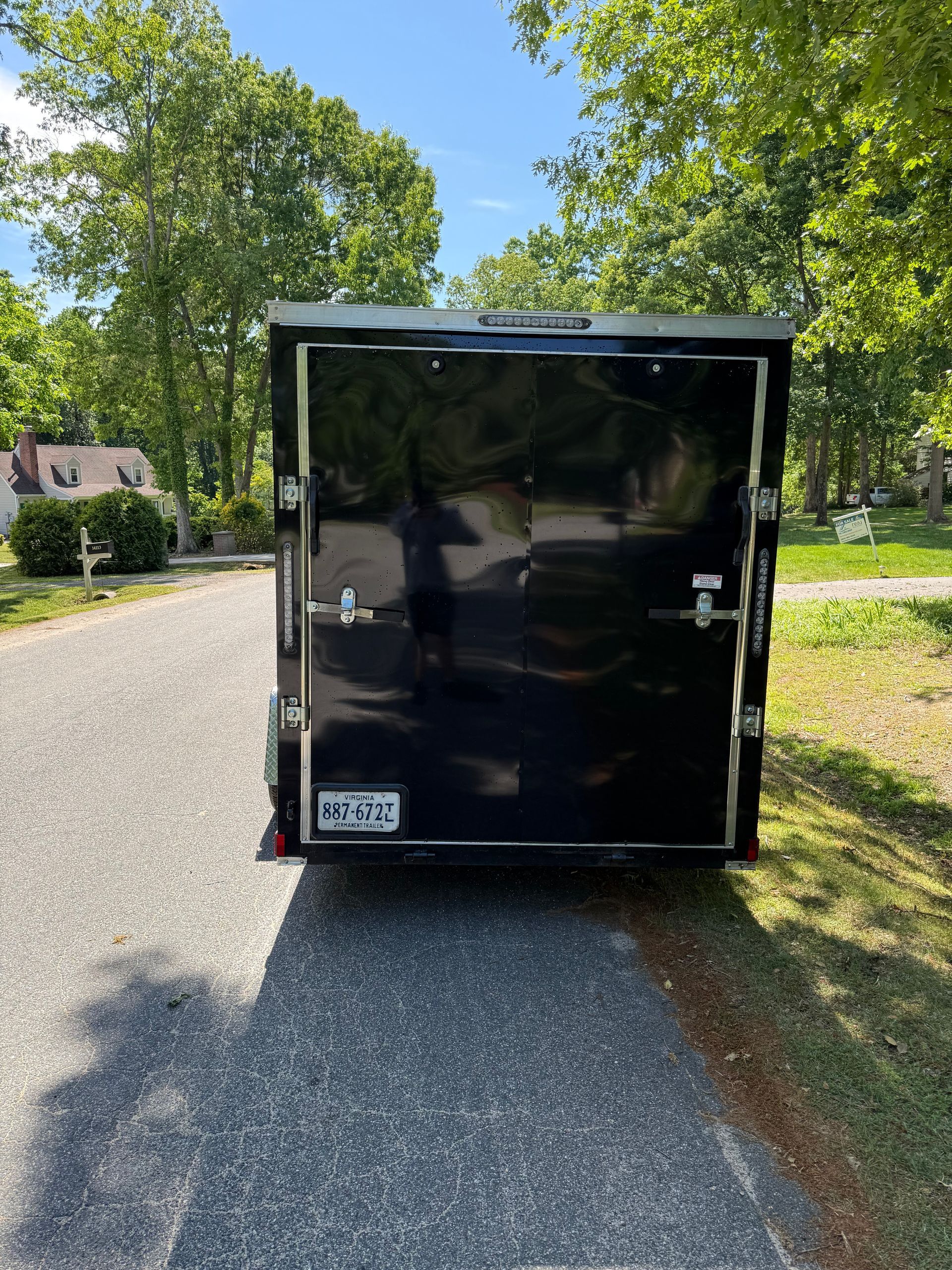Black cargo trailer parked on a gravel road beside trees and a house