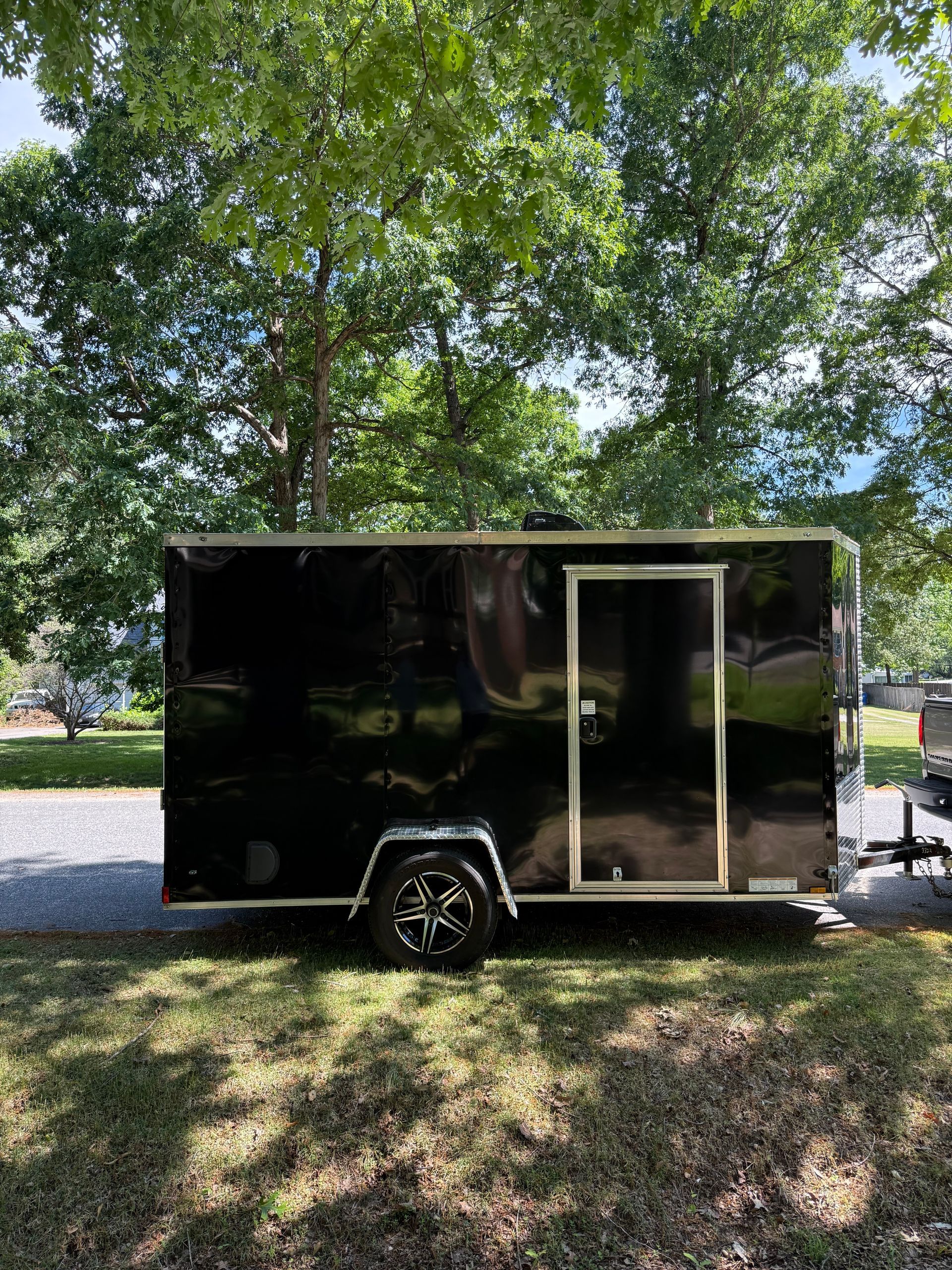 Black enclosed utility trailer parked on grass under trees, with a side door and wheel visible.