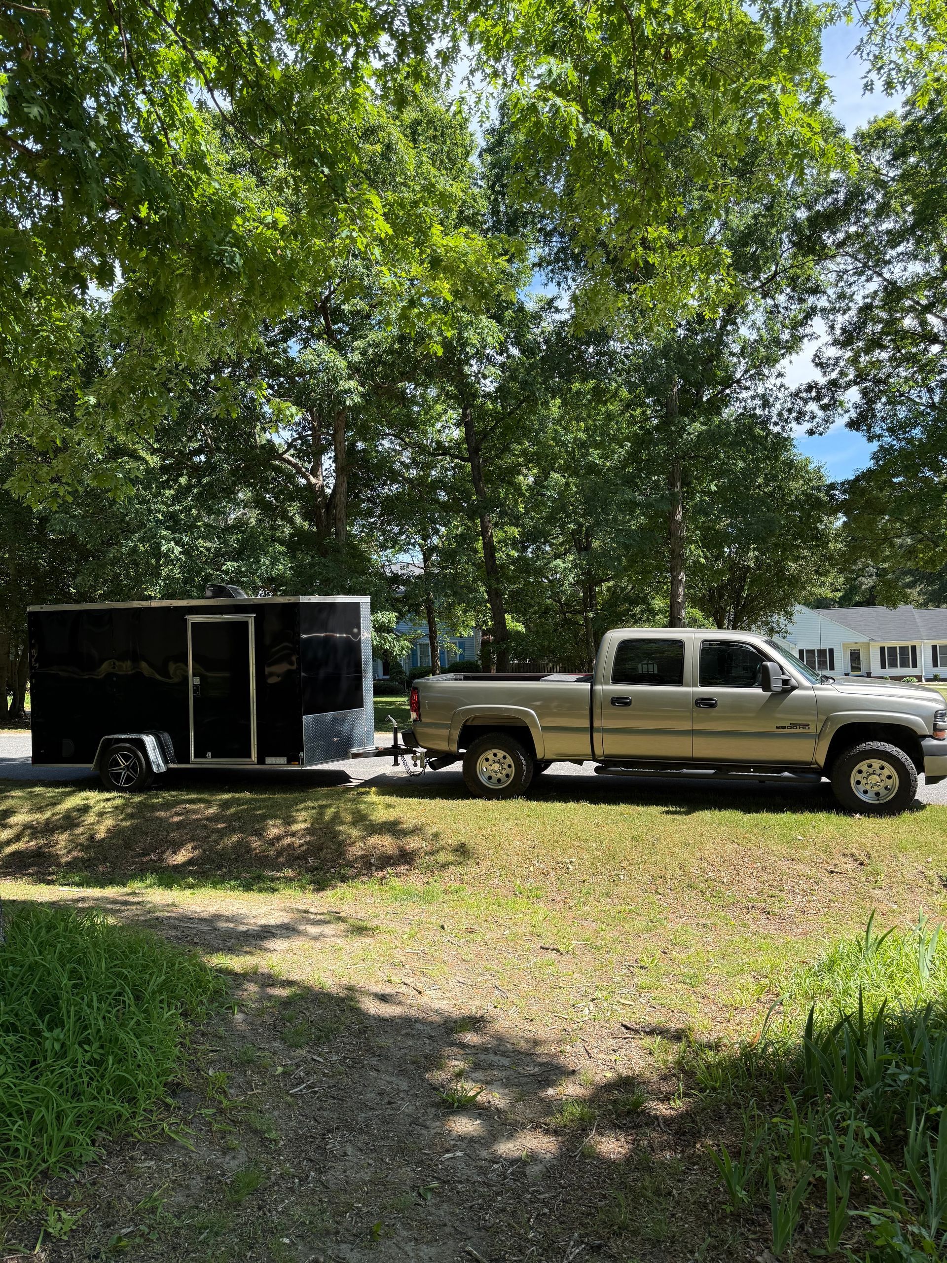 Silver pickup truck parked beside a black enclosed trailer under leafy trees in a yard.