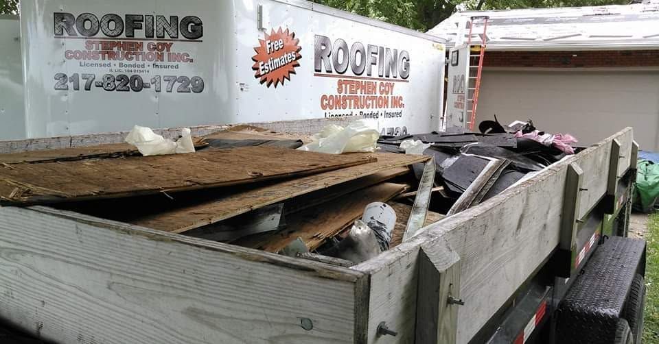 Flatbed trailer loaded with roofing debris and old shingles, parked beside a roofing company truck.