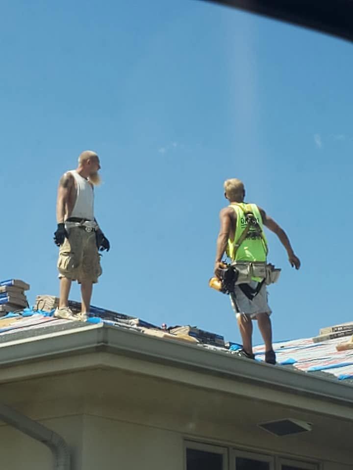 Two roofers in work gear stand on a house roof under a clear blue sky.