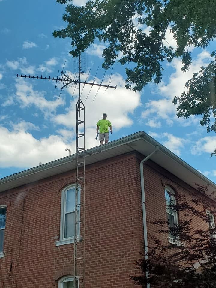 Worker in bright green gear standing on a brick house roof beside a tall antenna under a blue sky.