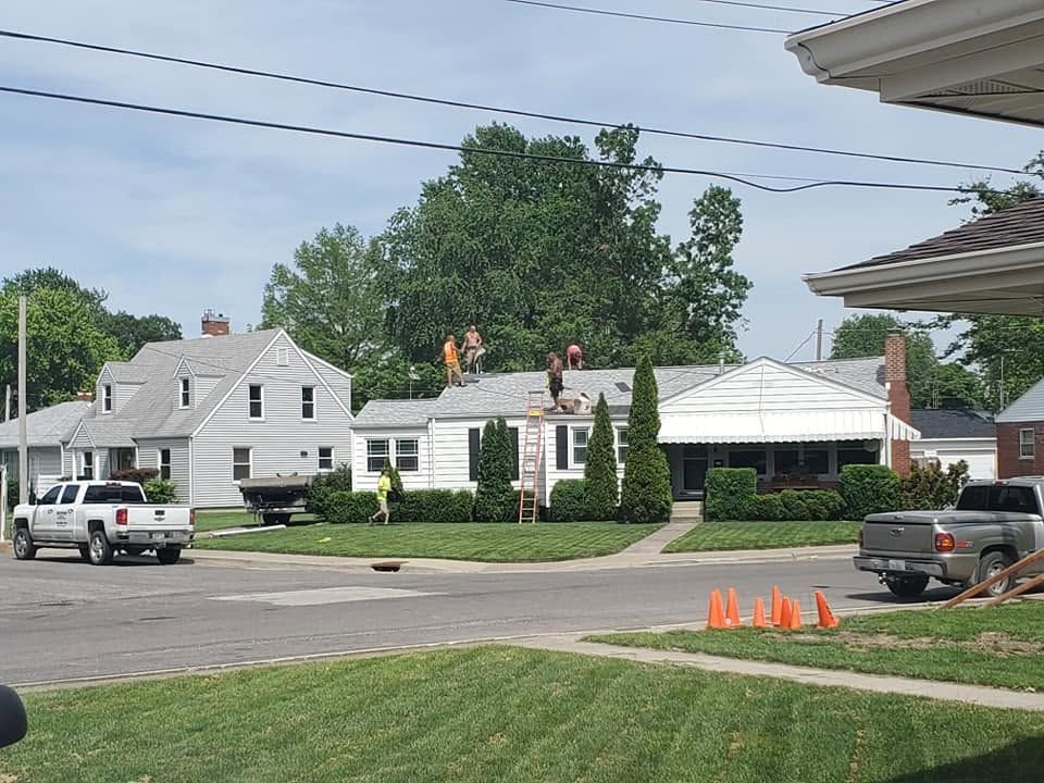 Workers on a white roof beside a house, with orange cones and parked vehicles in a suburban street.