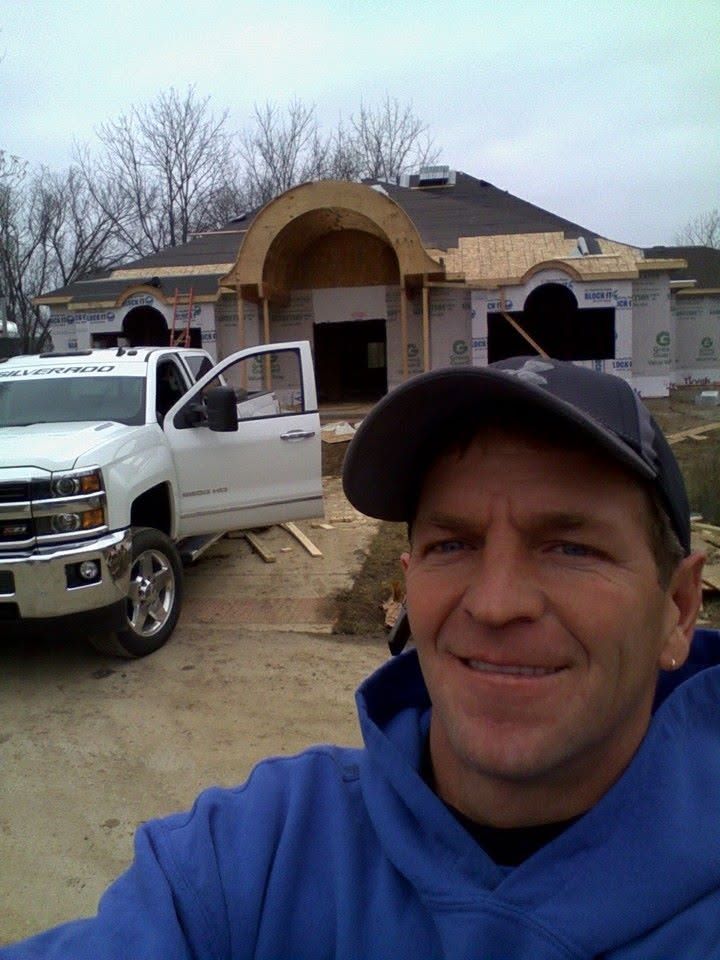 Man in blue hoodie and black cap taking a selfie in front of a house under construction, with a white truck nearby