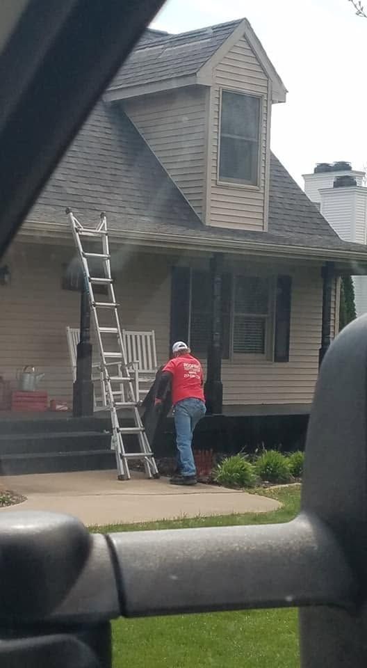 Two workers on a ladder repairing a house roof and porch, with a truck in the foreground.