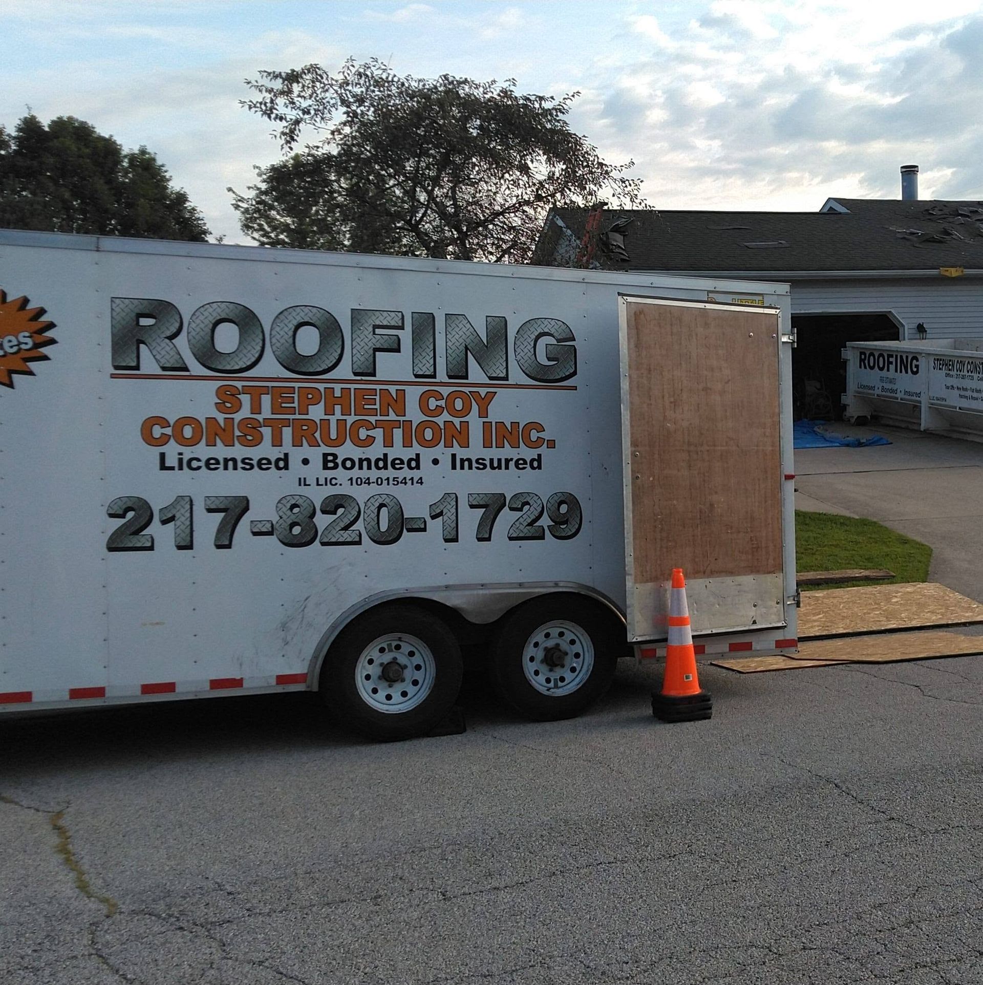 Roofing company trailer parked on a street beside a house, with orange traffic cones nearby.