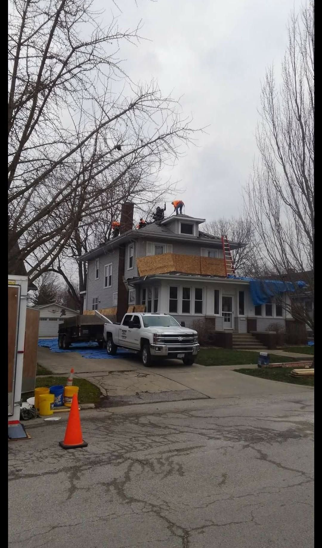 White two-story house with a parked van, orange cones, and leafless trees on a cloudy street