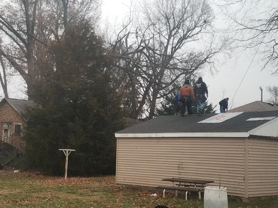 Workers on a shed roof repairing shingles beside a house and leafless trees