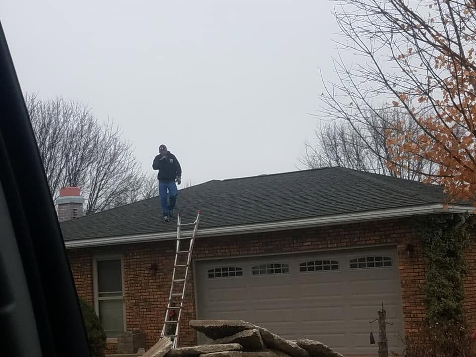 Person standing on a roof beside a ladder above a garage, with fallen tree limbs in the foreground