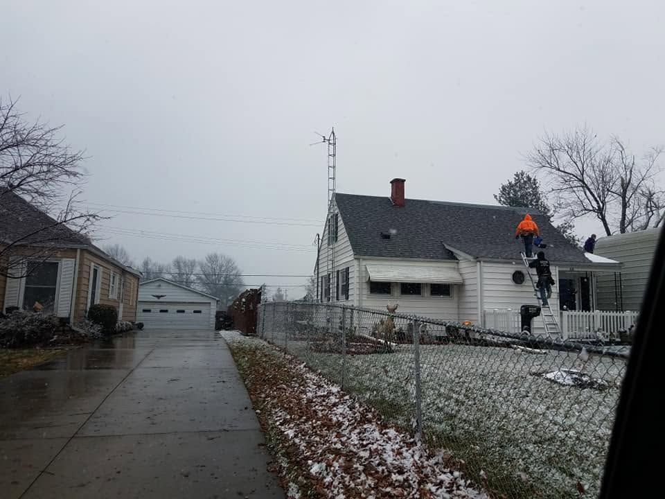 Snow-dusted suburban driveway beside a white house with an orange traffic cone on the roof and overcast sky