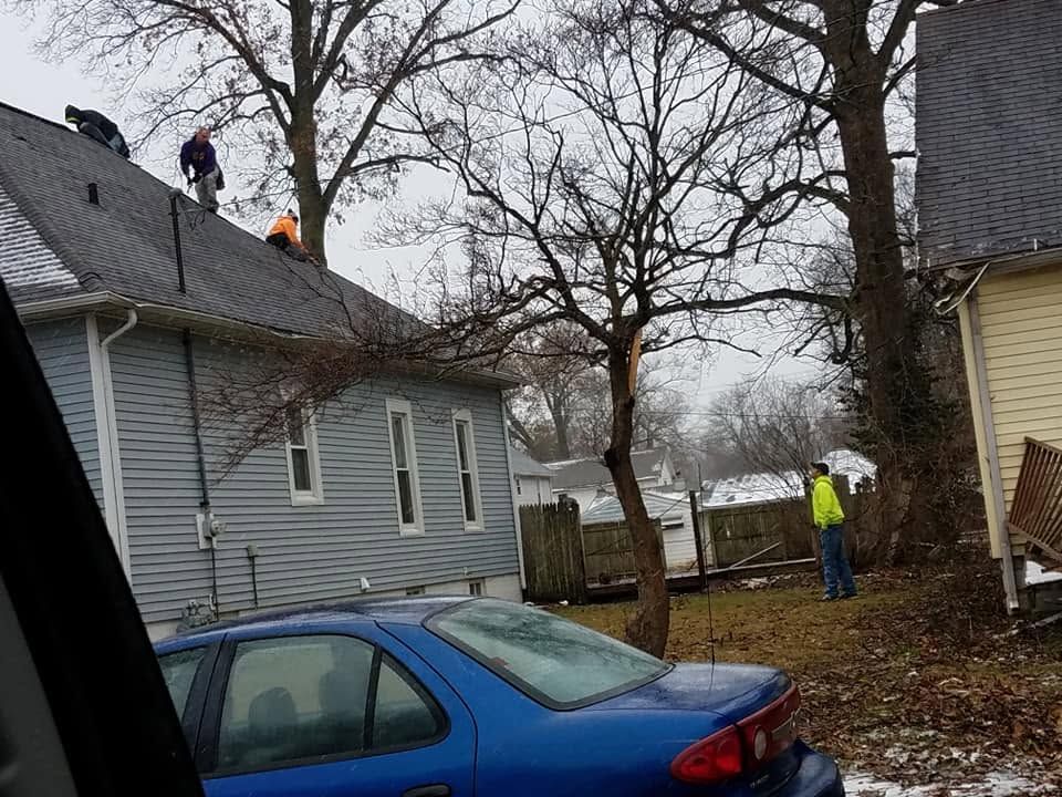 Workers on a gray house roof and in a yard beside a blue car on a cloudy day
