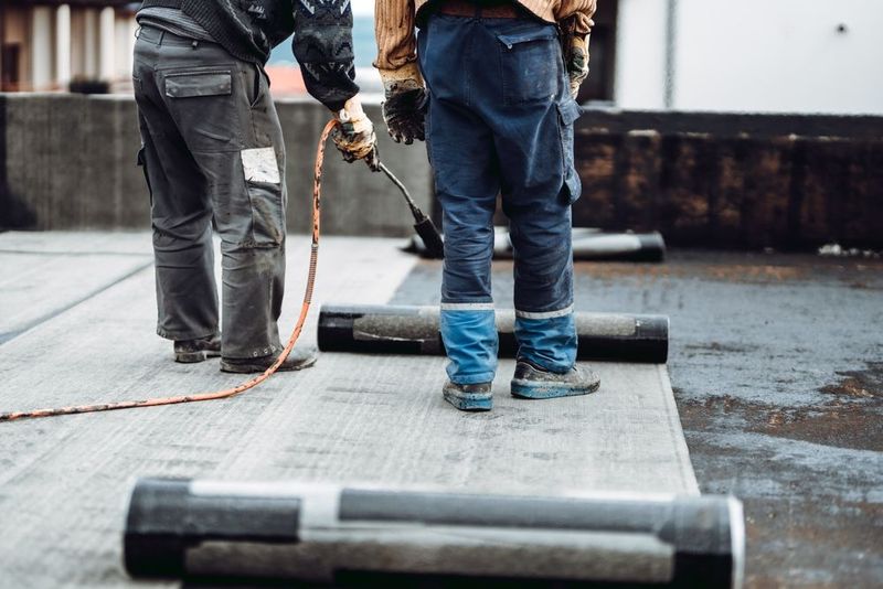 Two workers on a rooftop using a torch to seal roofing material on a wet surface