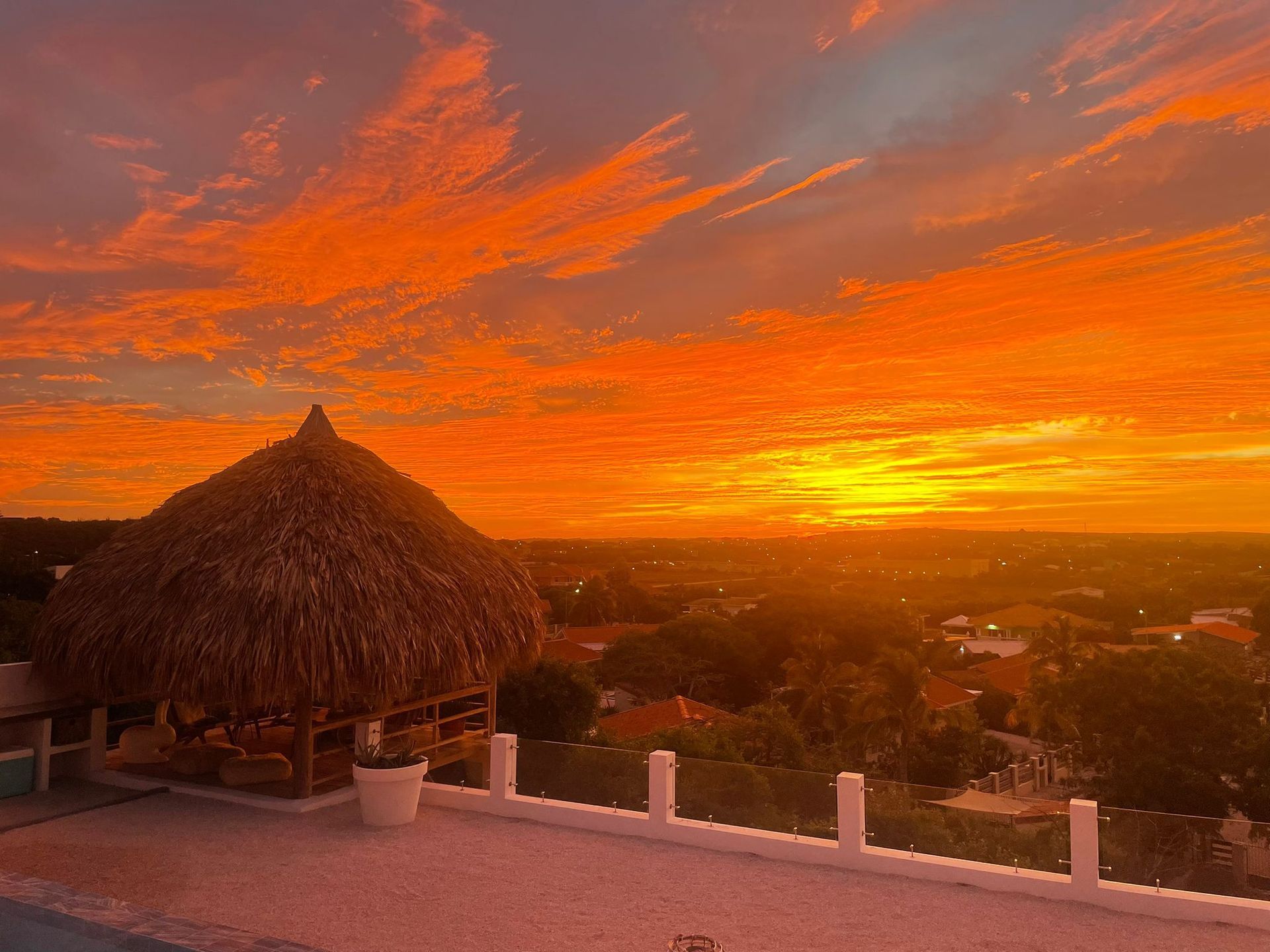 Zonsondergang boven een stad vanaf een dak, oranje lucht, hut met rieten dak, wit hek.