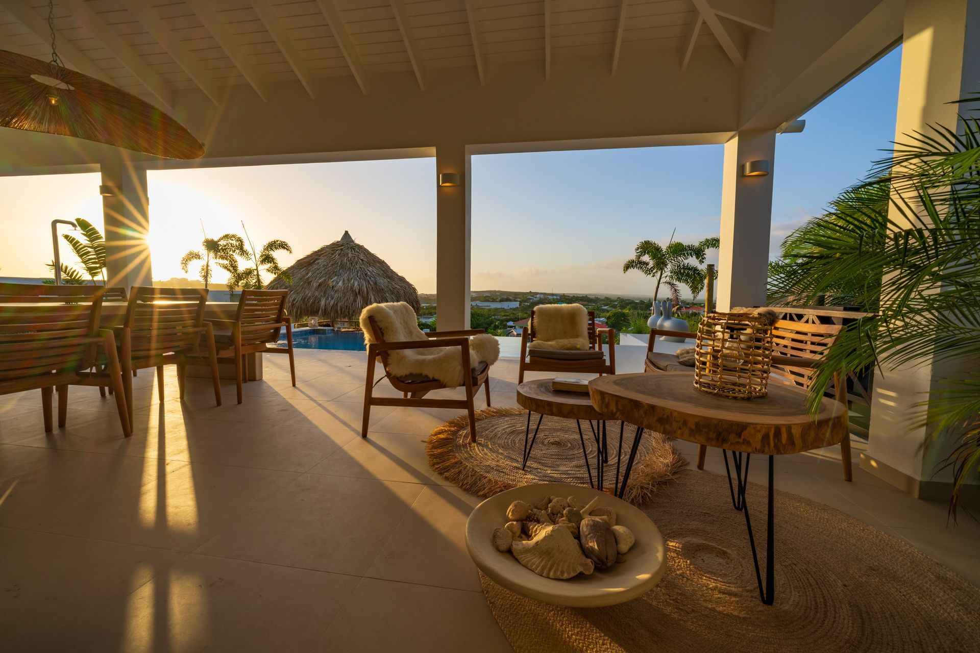 Overdekt terras met uitzicht op de zonsondergang, voorzien van stoelen, tafels en een zwembad.