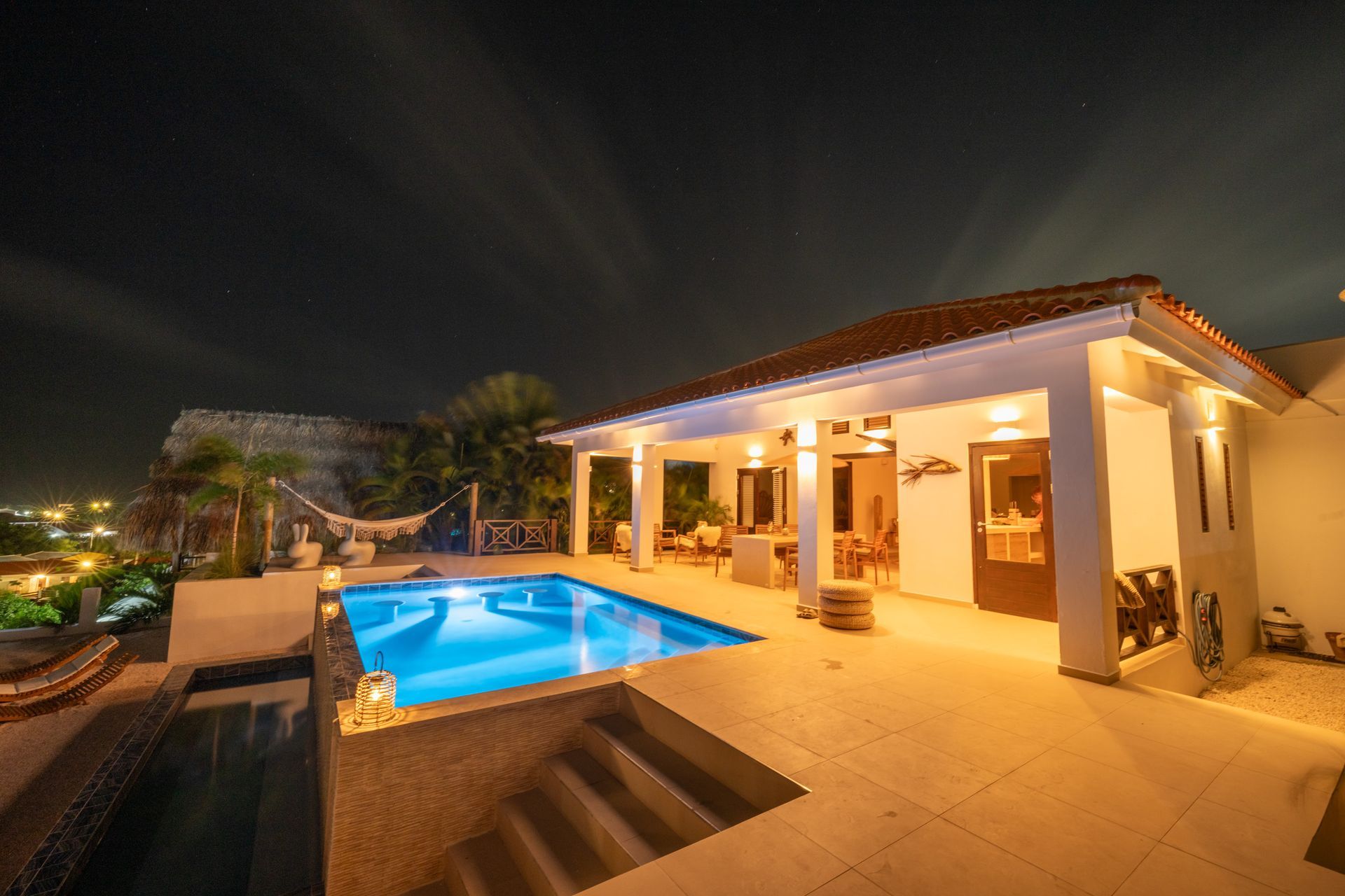 Nachtopname van een villa met een verlicht zwembad, terras en witte gevel onder een donkere hemel.
