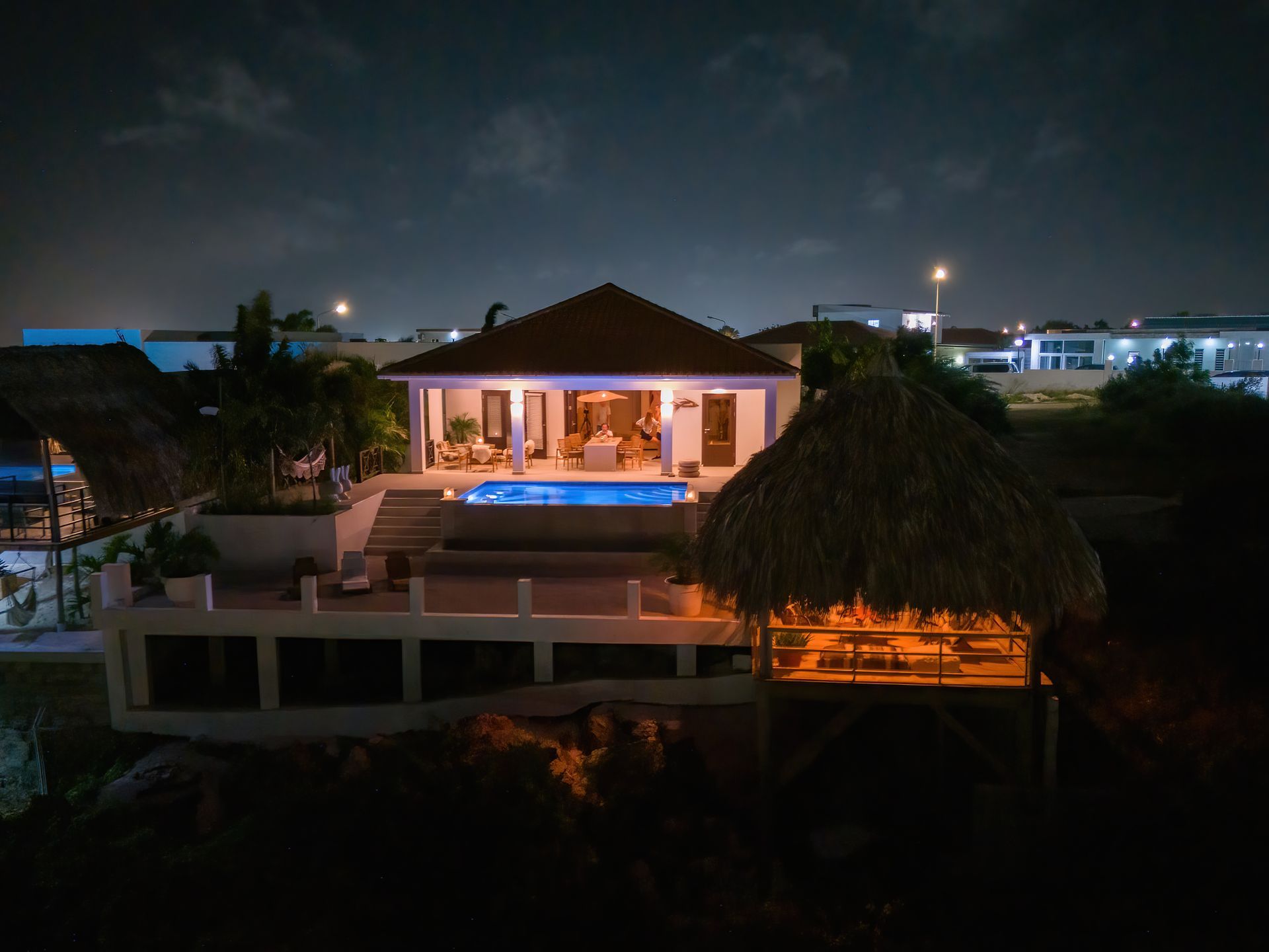 Nachtelijk uitzicht op een huis met een zwembad en rieten daken, alles verlicht.