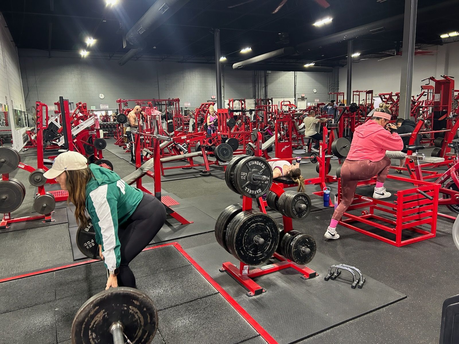 People working out at a busy, red-themed gym with various weight machines and equipment. Several individuals are lifting weights.