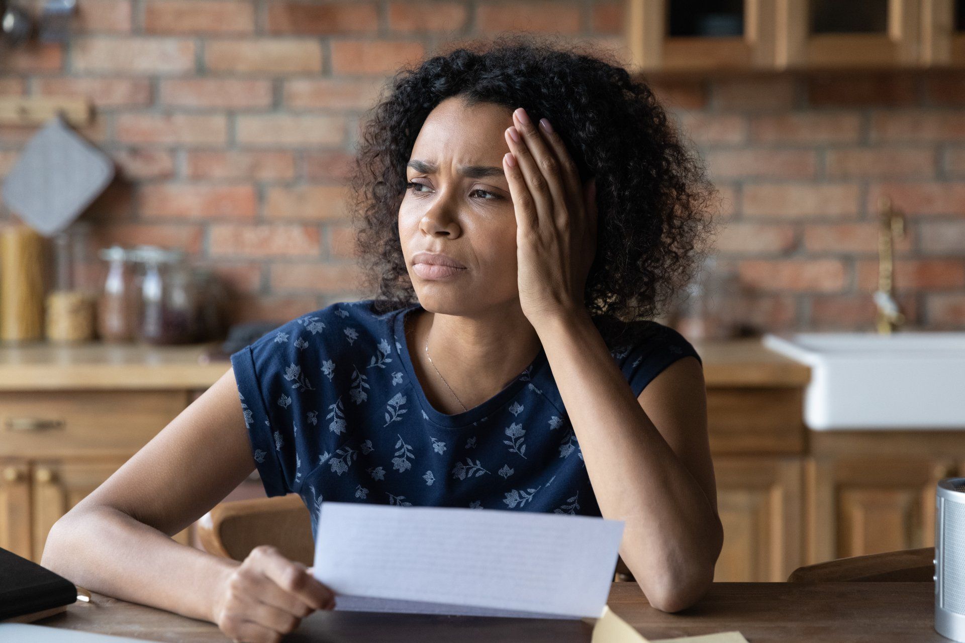 A woman sitting at a table holds a medical bill in her right hand, while her left hand touches the side of her face, she has a concerned expression.